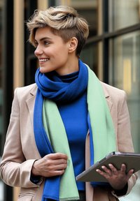 Person mit kurzen blonden Haaren, die ein beiges Jackett und einen zweifarbigen blau-grünen Schal trägt, hält ein Tablet in der Hand und lächelt, während sie nach links blickt.