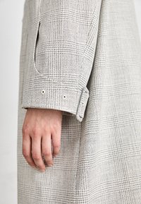 Lichtgrijze geruite jas van textuurstof. Heeft een manchet met een riemaccent en twee zichtbare knopen. Hand rustend op de mouw.