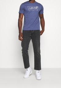 Modrá krátkosrstá trička s bílým logem Levi's na hrudi, v kombinaci s tmavě šedými rovnými džíny a bílými teniskami.