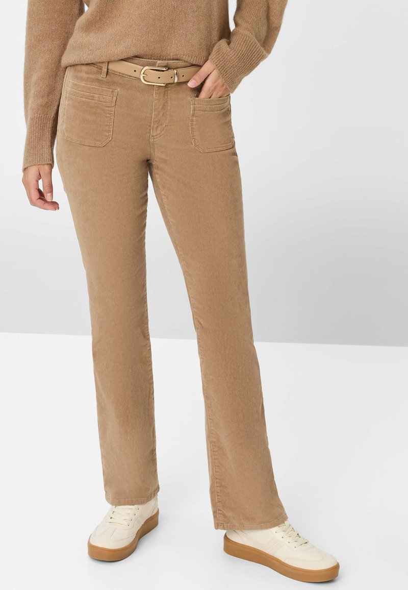 Kvinde iført beige corduroybukser med forreste lomme, et beige bælte, en brun sweater og cremede sneakers med gummisåler.