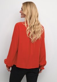 Red long-sleeve blouse with a relaxed fit, textured fabric, and gathered cuffs. Features a straight hem and a back yoke detail.