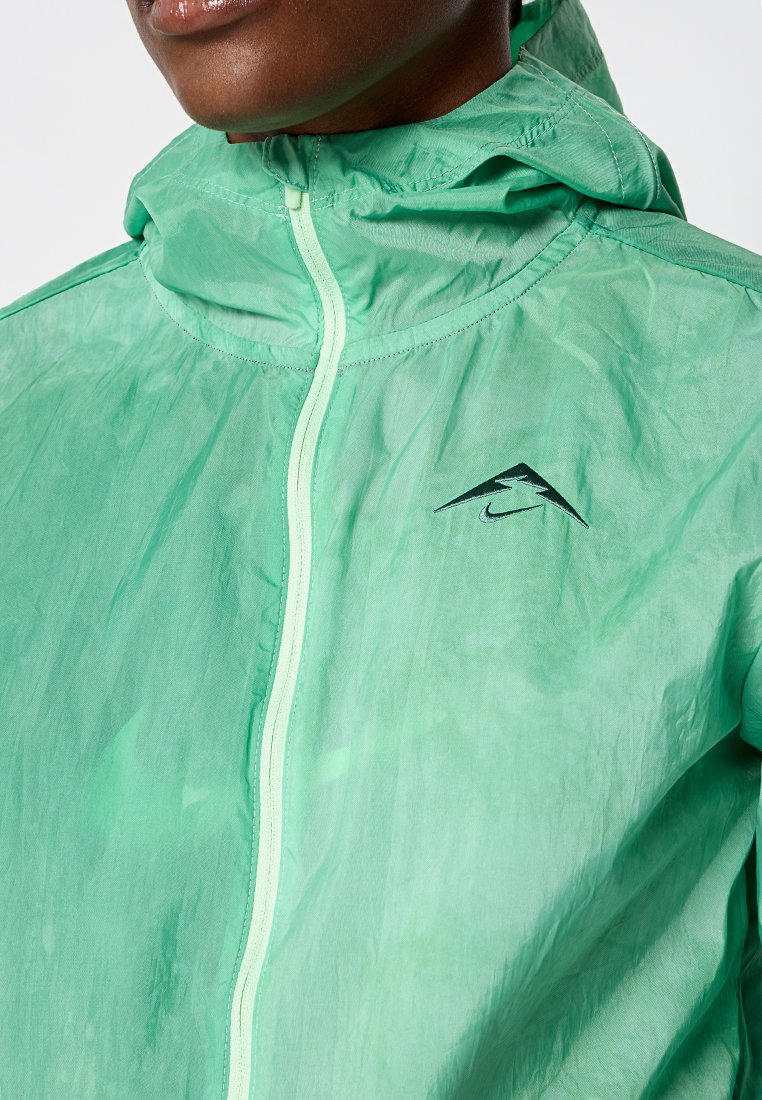 Lekka, półprzezroczysta zielona kurtka z kapturem, z centralnym zamkiem błyskawicznym i czarnym logo. Teksturowane wykończenie z subtelnymi wzorami.