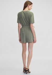 Olivgrüner Jumpsuit mit lockerer Passform, kurzen Ärmeln, einem elastischen Bund und zwei Gesäßtaschen. Kombiniert mit schwarzen Riemchenhighheels.