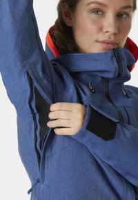 Blaue technische Jacke mit hohem Kragen, Reißverschluss, Netzbelüftungsöffnung unter dem Arm und kontrastierenden roten Akzenten an der Kapuze.