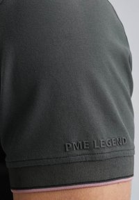 Polo shirt gris foncé avec un tissu texturé, logo noir "PME LEGEND" sur la manche et un accent rose contrastant sur le col.