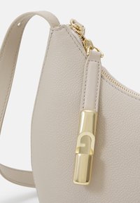 Borsa a mano in pelle beige con superficie texture, hardware della cerniera dorato e un distintivo accento dorato sul tirante della cerniera.