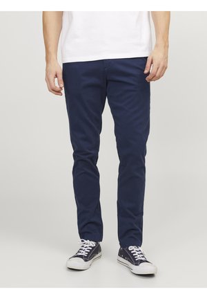Homme portant un pantalon chino bleu marine et des baskets basses noires avec des lacets blancs, se tenant devant un fond clair uni.