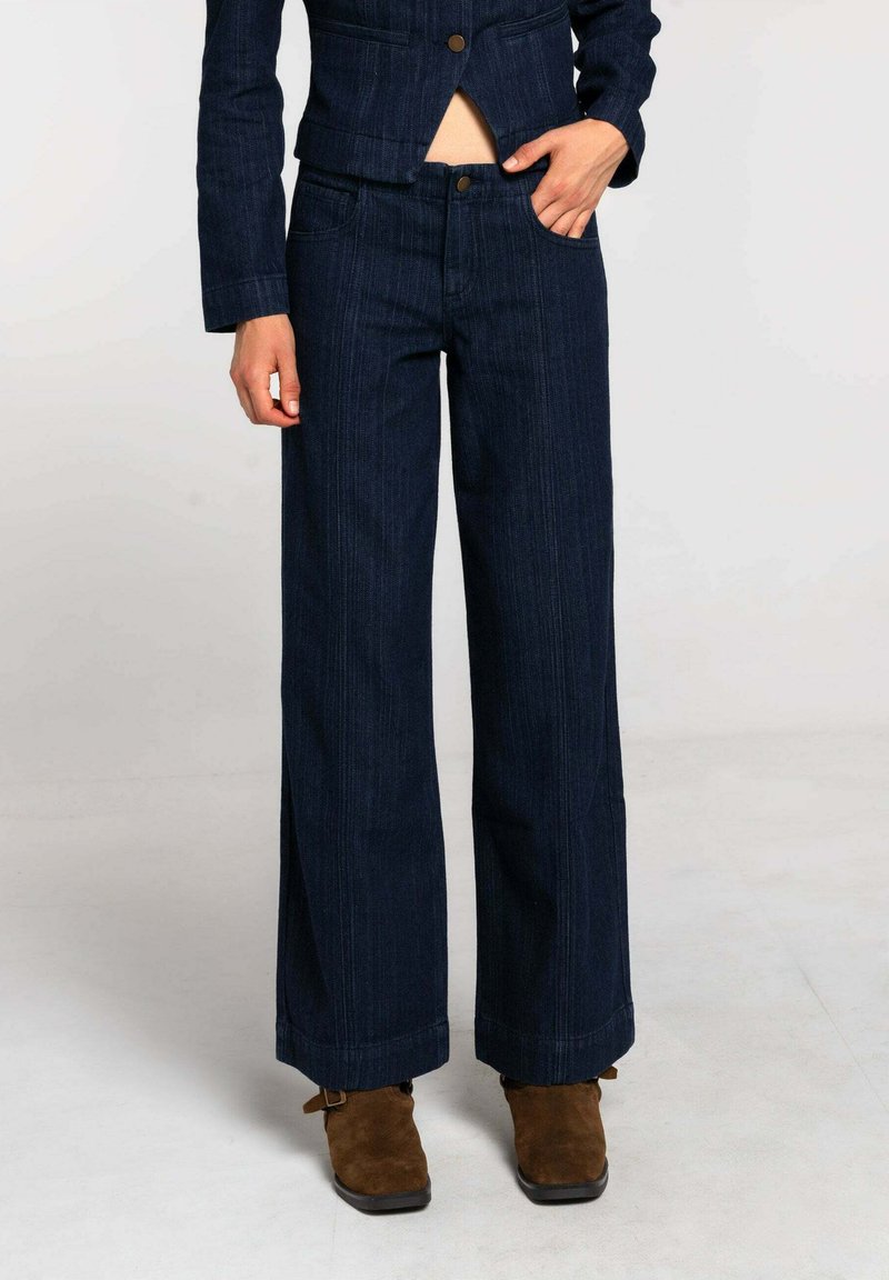 Pantalones anchos de mezclilla azul oscuro con cintura alta, costuras visibles, dos bolsillos frontales y cierre de botón. Tela texturizada y corte entallado.