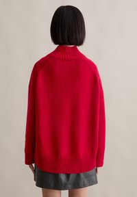 Roter oversized Pullover mit hohem Rippkragen, abgesenkten Schultern und Rippensaum, aus weichem, strukturiertem Strickstoff gefertigt.
