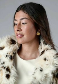 Cappotto in pelliccia finta con un motivo bianco e marrone, abbinato a un top crema. Orecchini a forma di foglia dorata aggiungono un tocco metallico. Texture liscia e naturale.