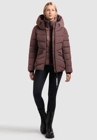 Brown puffer jacket with a high collar, zippered pockets, and hood. The fabric is quilted with a smooth texture. Worn with black leggings and boots.