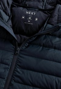 Svart quiltad jacka med dragkedja, med en mjuk innerfoder och etiketter som visar "vattentålig" och "värme". Storlek medium.