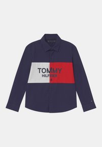 Niebieska koszula z długim rękawem z białym i czerwonym poziomym paskiem, na którym widnieje napis "TOMMY HILFIGER" w czerni. Standardowy kołnierzyk.