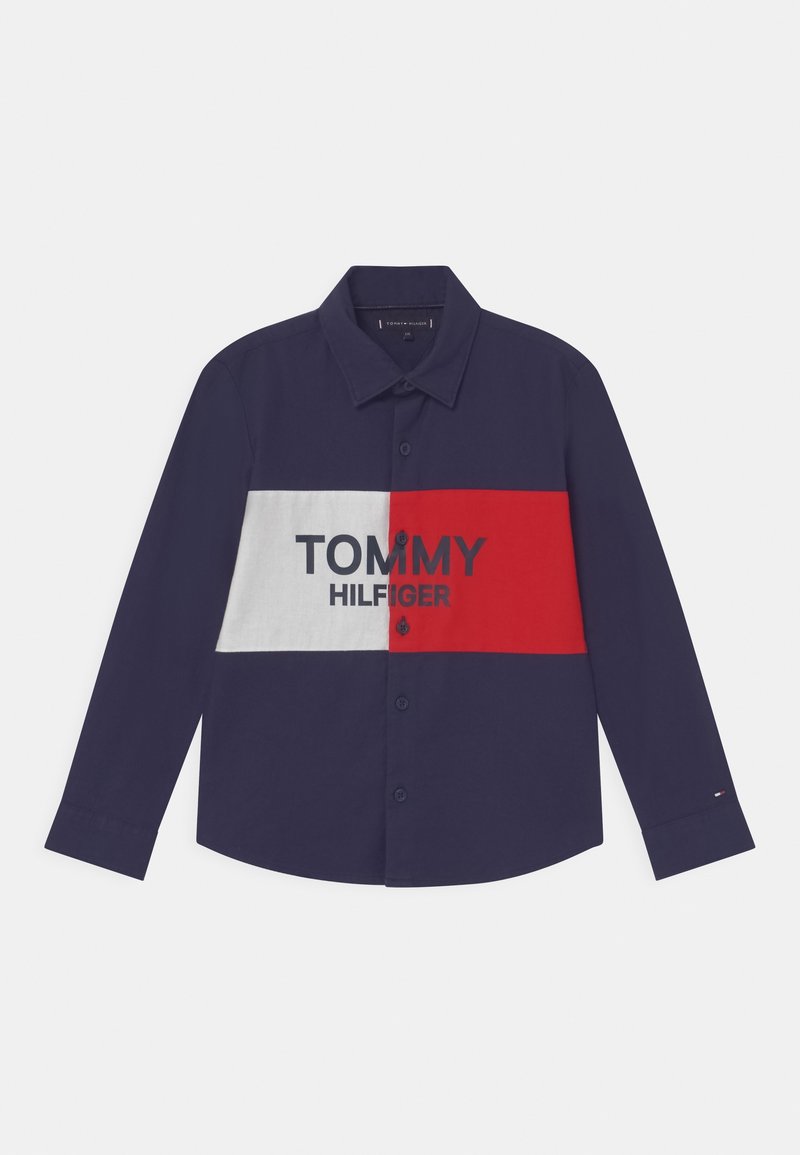 Niebieska koszula z długim rękawem z białym i czerwonym poziomym paskiem, na którym widnieje napis "TOMMY HILFIGER" w czerni. Standardowy kołnierzyk.