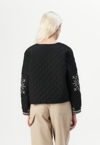 Black quilted jacket featuring decorative white embroidery on the sleeves and a round neckline. The design is boxy and cropped.