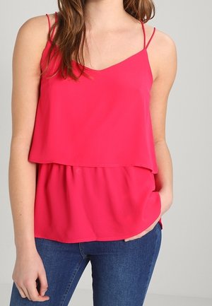 Frau trägt ein leuchtend pinkes ärmelloses, geschichtetes Oberteil mit dünnen Trägern, kombiniert mit blauen Jeans, Hand in der Tasche ruhend.