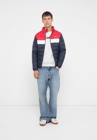 Polstret jakke med en rød, hvid og marineblå farveblokdesign, grå sweatshirt under, parret med lyseblå jeans og hvide sneakers.