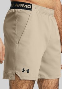 Beige Sportshorts mit einem elastischen Bund und einem schwarzen "ARMOR"-Logo. Enthält Seitentaschen und eine perforierte Textur am Saum.