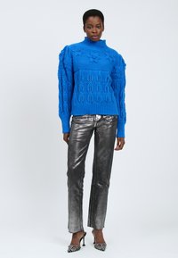 Maglione blu lavorato a maglia con motivi floreali e colletto alto, abbinato a pantaloni metallici argento e tacchi decorati. Sfondo piatto.