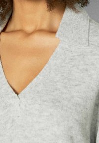 Harmaa neulottu neule, jossa on V-pääntie ja kaulus, esittelee pehmeää koostumusta ja huomaamatonta kuviota. Lähikuvassa kaula-aukon yksityiskohdat.