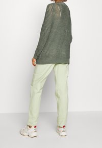 Osoba oblečená v volném tmavě zeleném pleteném svetru, světle zelených kalhotách a bílých teniskách New Balance, stojící na jednobarevném pozadí.