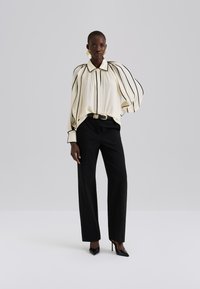 Κρεμ μπλούζα με κάθετες μαύρες ρίγες, ψηλό γιακά και μανίκια μπαλόνι, σε συνδυασμό με μαύρο παντελόνι και μαύρες μυτερές γόβες.