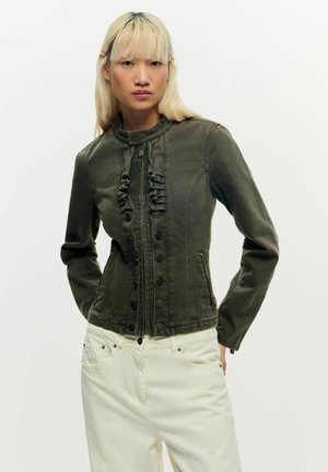 Jonge persoon met steil blond haar draagt donkergroene nauwsluitende jas met ruches en witte broek met hoge taille tegen een effen achtergrond.