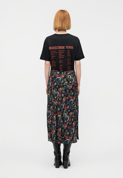 Μαύρο μπλουζάκι με κόκκινο γραφικό κείμενο στην πλάτη, συνδυασμένο με μακριά φλοράλ φούστα σε χρώματα όπως κόκκινο, μπλε και πράσινο, φορεμένο με μαύρες μπότες.