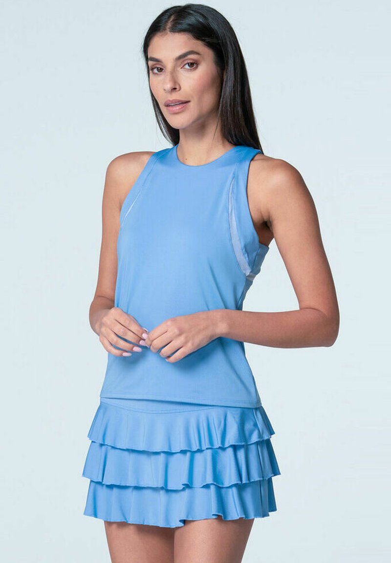 Mujer con cabello largo y oscuro que lleva una blusa sin mangas de color azul claro y una falda de volantes a juego, de pie frente a un fondo liso.