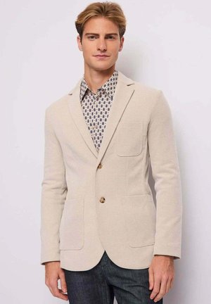 Giovane uomo con capelli corti biondi che indossa un blazer beige sopra una camicia a fantasia abbottonata e jeans scuri, in piedi davanti a uno sfondo semplice.