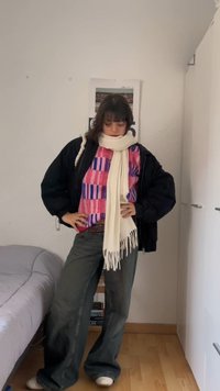 Άτομο που στέκεται σε εσωτερικό χώρο φορώντας μαύρο μπουφάν, πολύχρωμη μπλούζα με σχέδια, λευκό κασκόλ, παντελόνι με φαρδιά πόδια, με τα χέρια στη μέση σε ένα υπνοδωμάτιο.