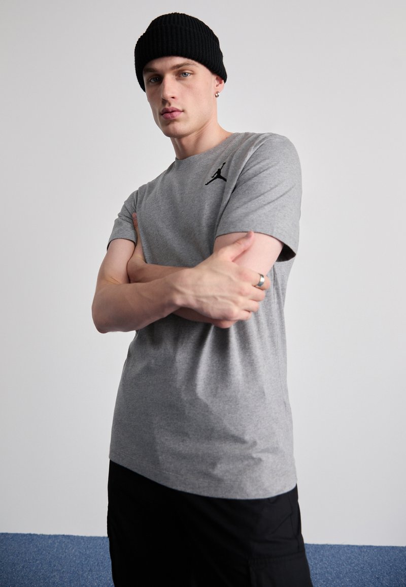 Tricou gri cu mânecă scurtă, având un logo negru Jordan pe pieptul stâng, purtat sub o căciulă neagră din tricot. Material din bumbac neted.