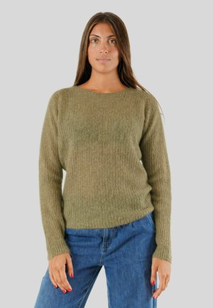 Donna che indossa un maglione di lana verde oliva e jeans blu, in piedi davanti a uno sfondo grigio chiaro uniforme.