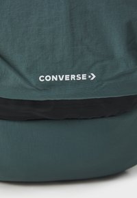 Grön Converse-ryggsäck i ett texturerat material, med en svart meshficka och vitt logotryck på framsidan.