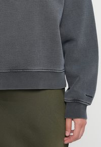 Gray sweatshirt with ribbed cuffs and hem, soft fabric texture, featuring a logo on the sleeve, paired with an olive green lower garment.