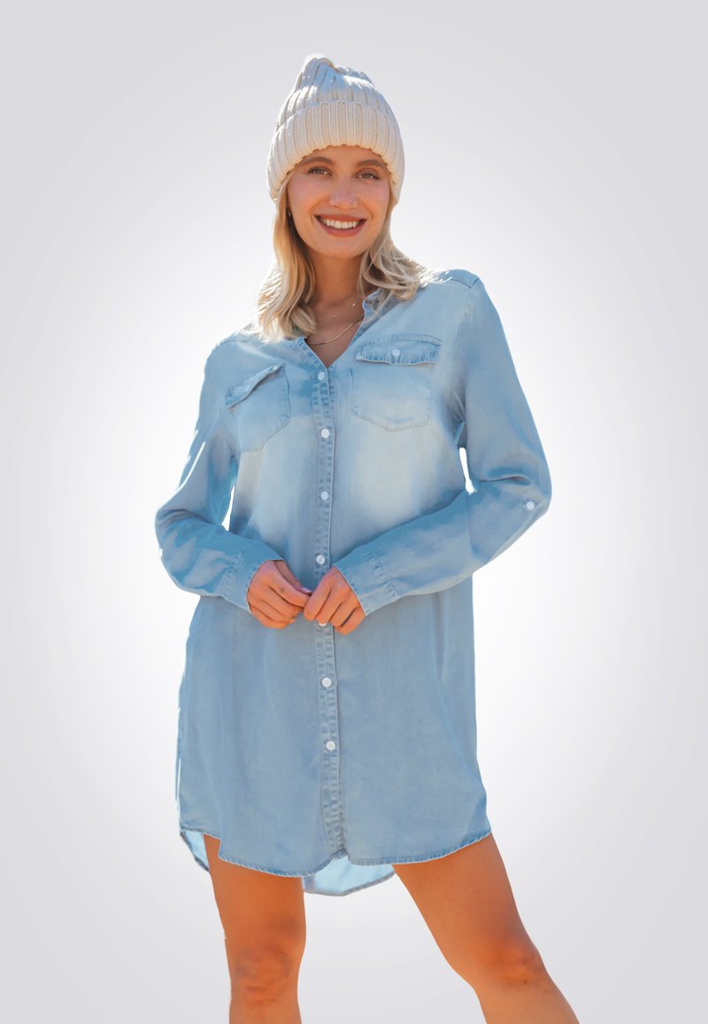 Vestido camisa de mezclilla azul claro con mangas largas, cierre de botones en la parte frontal, dos bolsillos en el pecho y un dobladillo redondeado. Combinado con un gorro de punto color crema.