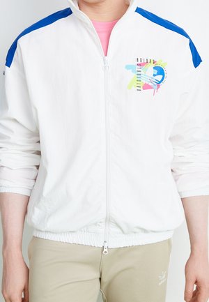 Persoon draagt een wit jack met ritssluiting en blauwe schouderpanelen, met een kleurrijk Adidas Originals-logo, gecombineerd met beige broek.