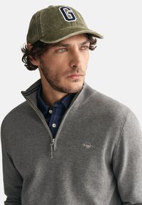 Grauer Zip-Pullover mit geripptem Kragen, getragen über einem blauen Karoshemd. Die olivgrüne Mütze hat ein hervortretendes weißes "G"-Logo.