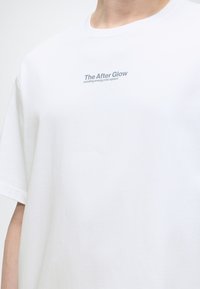 Άτομο που φοράει λευκό μπλουζάκι με το κείμενο «The After Glow στέλνει ενέργεια στο διάστημα» τυπωμένο στο στήθος.