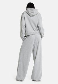 Άτομο που στέκεται με την πλάτη, φοράει φαρδιά ανοιχτογκρί φούτερ με κουκούλα και φαρδιά φόρμα, σε λευκό απλό φόντο.