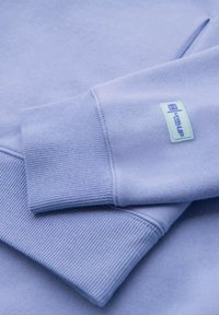 Gros plan sur un sweat-shirt violet clair en tissu mélangé coton, avec des poignets côtelés et une petite étiquette logo brodée en vert pâle. Texture lisse dans l'ensemble.