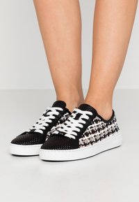 Jambes portant des baskets en cuir tressé noir et blanc avec des lacets blancs et des semelles épaisses blanches sur un fond clair uni.