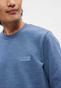 Sweatshirt azul com decote redondo e tecido texturizado, apresentando um rótulo cosido de cor azul clara no lado esquerdo do peito.