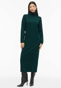 Tumši zaļš turtlenecks kleita, garas piedurknes, brīvs griezums, ribots tekstūra, līdz ceļiem garš, apvienota ar melniem augstpumpuriem zābakiem.