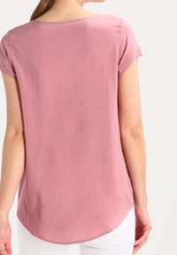 Roze top met korte mouwen, een gladde textuur, een ronde halslijn en een afgeronde zoom, van achteren getoond tegen een neutrale achtergrond.