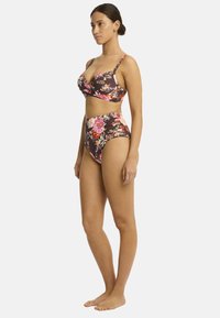 La modella posa di lato, indossando un bikini floreale su uno sfondo scuro. La parte superiore ha spalline sottili e i pantaloni a vita alta esaltano la sua figura.