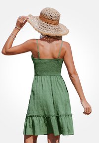 Vestito verde senza maniche con corpetto arricciato e orlo a balze sovrapposte. Abbinato a un cappello di paglia a tesa larga, che mostra un tessuto strutturato.