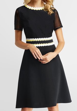 Black knee-length dress with mesh short sleeves, scalloped white and yellow trim at neckline and waist, worn by a standing woman.
