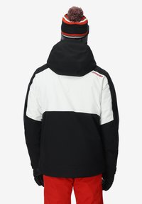 Giacca insulated in bianco e nero con cappuccio, caratterizzata da un logo rosso. Abbinata a pantaloni rossi e un berretto nero con pompon rosso.