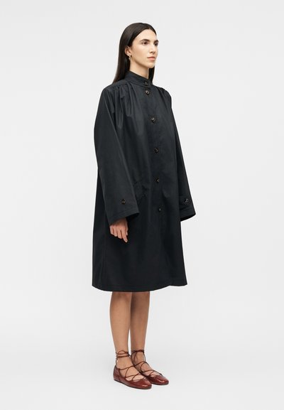 Femme debout de face portant un long manteau noir ample à boutons avec des manches larges et des ballerines marron à lacets, sur un fond uni.